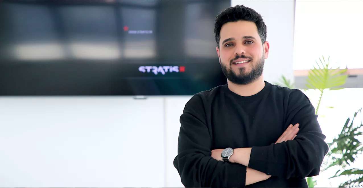 Un homme souriant avec une barbe courte et des cheveux foncés bouclés pose les bras croisés dans un bureau moderne et lumineux. Il porte un pull noir et une montre argentée à son poignet gauche. Derrière lui, un grand écran noir affiche le logo 'STRATIS' et un mur blanc avec des fenêtres qui laissent entrer la lumière naturelle. Quelques plantes vertes apportent une touche de fraîcheur à l’arrière-plan.