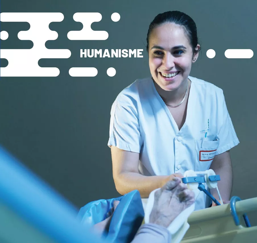 Une soignante en blouse blanche, arborant un large sourire, interagit avec bienveillance avec un patient alité dont la main fragile repose sur la barrière du lit, dans une atmosphère douce et chaleureuse, illustrant le mot « HUMANISME » inscrit en haut à gauche de l’image.