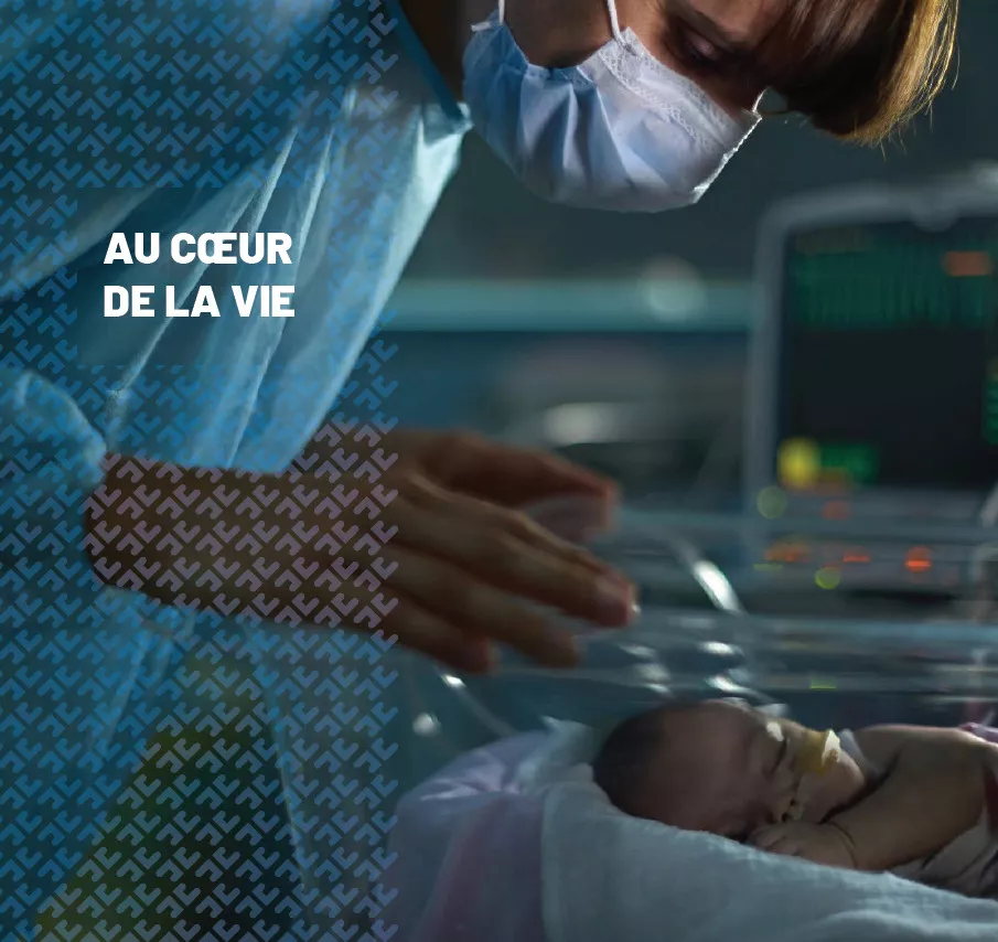 Une soignante en blouse bleue et masque chirurgical se penche avec attention vers un nouveau-né prématuré allongé dans une couveuse, sous un éclairage tamisé, avec un moniteur médical affichant des constantes vitales en arrière-plan ; le texte « AU CŒUR DE LA VIE » est inscrit sur la gauche de l’image.