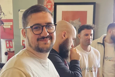 Un homme souriant avec des lunettes rondes et un sweat clair est au premier plan, tandis qu’en arrière-plan, plusieurs personnes discutent dans une pièce décorée. - Agrandir l'image 2 sur 28, fenêtre modale