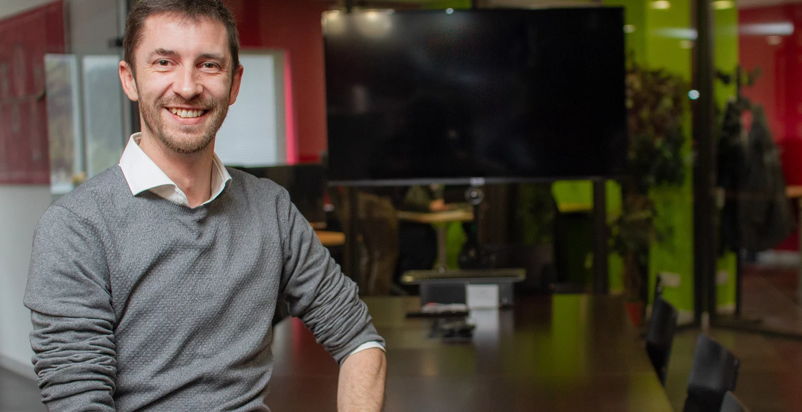 Un homme souriant avec une barbe courte et des cheveux bruns, portant une chemise blanche sous un pull gris, est assis dans une salle de réunion moderne. Il est appuyé sur une table de conférence sombre, avec un grand écran noir et des chaises autour. L'arrière-plan montre un bureau avec des murs colorés en rouge et vert, ainsi que des plantes et des employés travaillant à leurs postes.