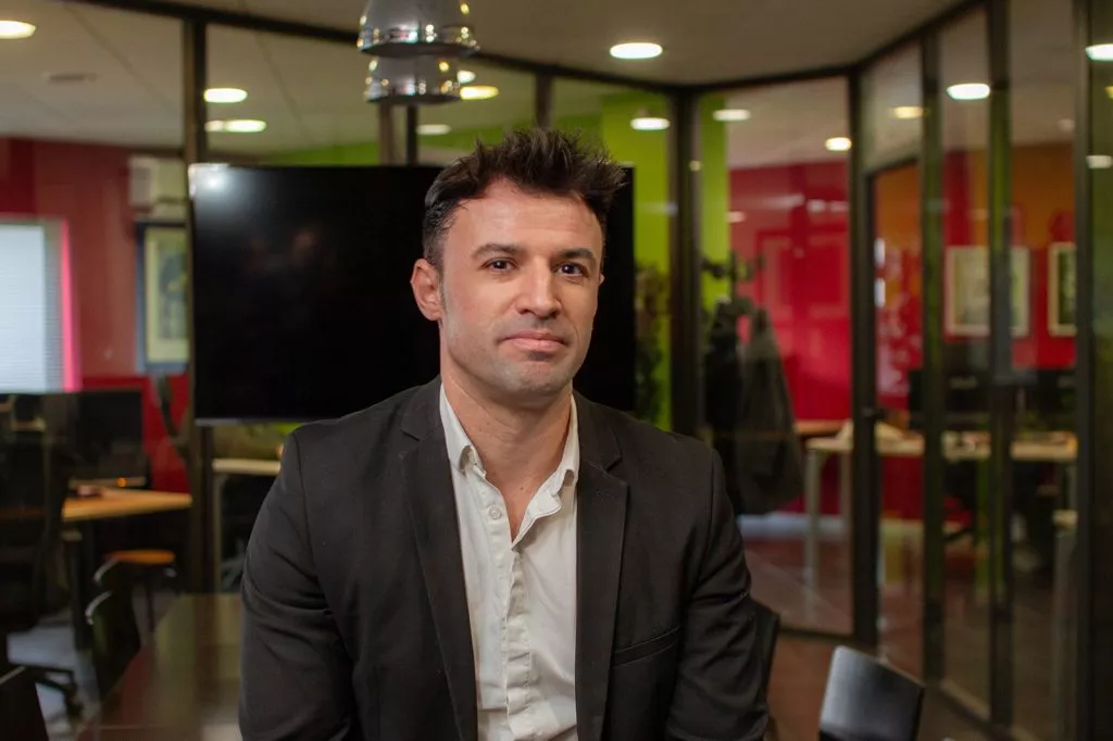 Un homme aux cheveux courts et bruns, habillé d’un costume noir et d’une chemise blanche, est assis dans un bureau moderne entouré de parois vitrées avec des murs colorés en rouge et vert, et un écran noir en arrière-plan.