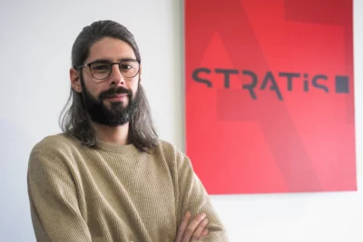 Un homme aux cheveux longs, à la barbe fournie et portant des lunettes rondes ainsi qu’un pull beige, pose les bras croisés avec un léger sourire devant un mur blanc orné d’un grand panneau rouge affichant le logo "STRATIS". - Agrandir l'image 8 sur 12, fenêtre modale