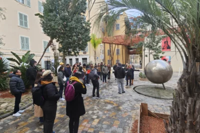 Un groupe de personnes, habillées chaudement, se rassemble dans une jolie place pavée entourée de bâtiments aux façades claires et de végétation, écoutant attentivement un guide qui semble leur donner des explications, près d’une sculpture sphérique en métal représentant un globe terrestre. - Agrandir l'image 12 sur 28, fenêtre modale