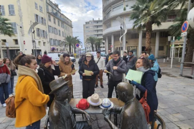 Un groupe de personnes, tenant des feuilles en main, participe à une activité en plein air autour d’une sculpture en bronze représentant deux hommes assis à une table de jeu, sur laquelle plusieurs chapeaux sont posés, dans une place animée bordée de palmiers et de bâtiments à l’architecture méditerranéenne. - Agrandir l'image 13 sur 28, fenêtre modale