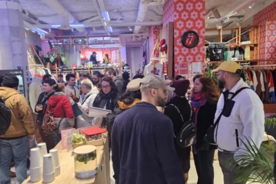 Une foule de personnes, vêtues de manteaux et de bonnets, se rassemble dans une boutique au décor rétro avec des murs recouverts de papier peint à motifs fleuris, des étagères remplies de vêtements et d’accessoires, ainsi qu’un coin boissons avec des gobelets empilés et un bocal d’infusion sur une table. - Agrandir l'image 4 sur 28, fenêtre modale