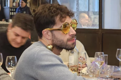 Un homme portant un pull bleu est assis à une table dans un restaurant animé, affichant un style humoristique avec des lunettes rondes teintées dorées et des boules de Noël accrochées aux oreilles en guise de boucles d’oreilles, tandis que les autres convives profitent de la soirée dans une ambiance chaleureuse et conviviale. - Agrandir l'image 7 sur 28, fenêtre modale