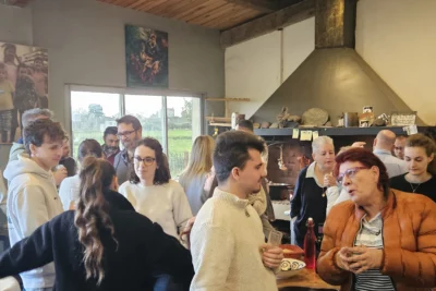 Un groupe de personnes échange dans une ambiance chaleureuse et conviviale autour d’un grand foyer en pierre, dans un intérieur rustique décoré de photos et d’objets anciens, tandis qu’une femme en doudoune orange semble raconter une histoire animée à ses interlocuteurs. - Agrandir l'image 15 sur 28, fenêtre modale