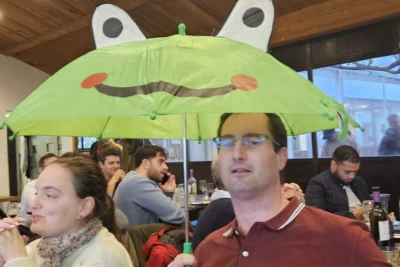 Un homme assis à table dans un restaurant tient avec sérieux un parapluie vert en forme de grenouille, avec un large sourire dessiné et des oreilles en relief, créant un contraste humoristique avec l’ambiance du repas et l'attitude plus détendue des autres convives en arrière-plan. - Agrandir l'image 9 sur 28, fenêtre modale