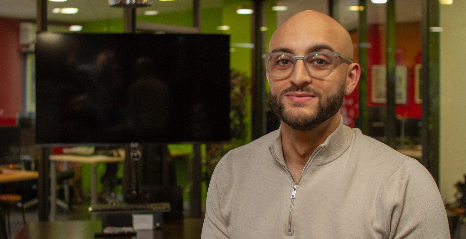 Un homme chauve avec une barbe bien taillée et des lunettes transparentes pose dans une salle de réunion moderne. Il porte un pull beige avec un col zippé et arbore un léger sourire. L’arrière-plan montre un espace de travail avec des parois vitrées, un grand écran noir, des bureaux et des murs colorés en rouge et vert, créant une atmosphère dynamique et professionnelle.