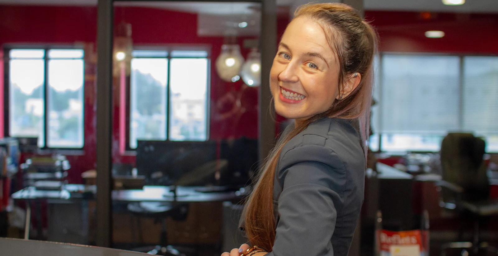 Une femme souriante aux longs cheveux bruns attachés en arrière pose avec une expression joyeuse dans un bureau moderne. Elle porte une veste sombre et semble tournée vers la caméra avec une posture dynamique et accueillante. L’arrière-plan, visible à travers une paroi vitrée, montre un espace de travail lumineux avec des bureaux, des chaises et des lampes suspendues. Les murs rouges et les grandes fenêtres laissent entrer la lumière naturelle, créant une ambiance chaleureuse.
