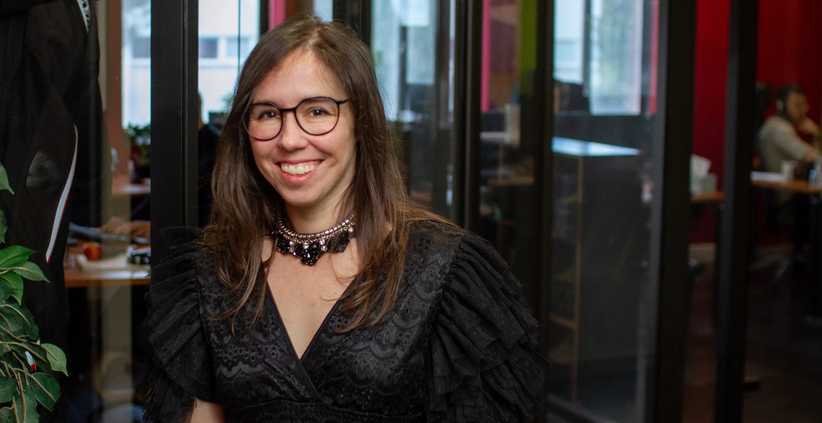 Une femme souriante avec des lunettes et des cheveux longs bruns pose dans un bureau moderne. Elle porte une robe noire élégante avec des manches en dentelle volumineuses et un collier orné de pierres noires. Derrière elle, une paroi vitrée sépare l’espace, laissant entrevoir plusieurs bureaux avec des employés en train de travailler. L’environnement est lumineux, avec des murs colorés en rouge et vert, ainsi que des plantes décoratives.