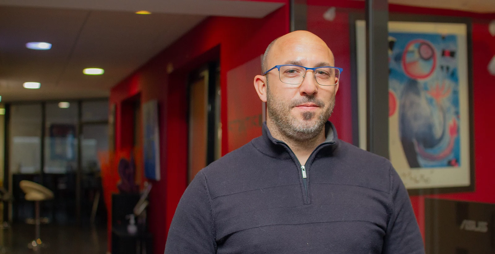 Un homme chauve avec une barbe courte et des lunettes à monture bleue pose dans un espace moderne. Il porte un pull noir avec un col zippé et affiche une expression confiante et sereine. L’arrière-plan présente un couloir avec des murs rouges, des vitres donnant sur des bureaux, et une œuvre d’art abstraite encadrée sur le mur. L’éclairage tamisé et les éléments de mobilier modernes créent une ambiance professionnelle et chaleureuse.