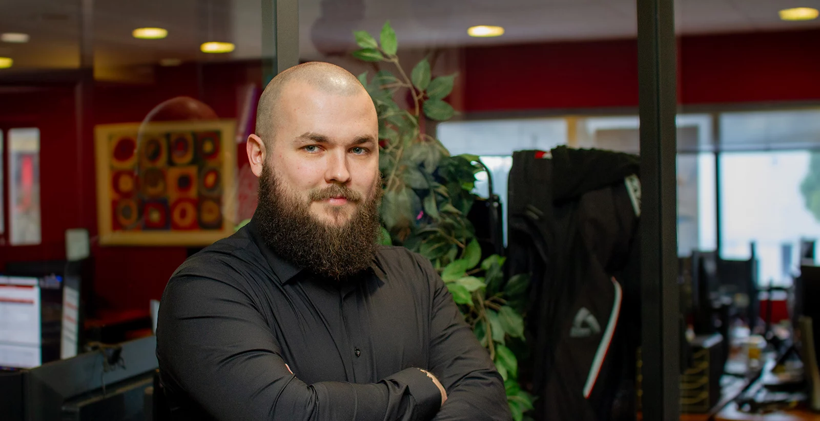 Un homme chauve avec une barbe longue et fournie pose les bras croisés dans un bureau moderne. Il porte une chemise noire et arbore un regard confiant. L’arrière-plan montre un espace de travail avec des bureaux, des écrans d’ordinateur, une plante verte et un mur rouge décoré d’une œuvre d’art abstraite encadrée. La lumière naturelle entre par de grandes fenêtres, ajoutant une ambiance chaleureuse à la scène