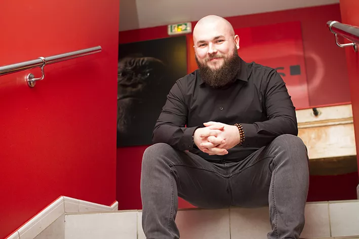 Un homme barbu, souriant et vêtu de noir est assis sur les marches d’un escalier entouré de murs rouges, avec un décor moderne en arrière-plan.