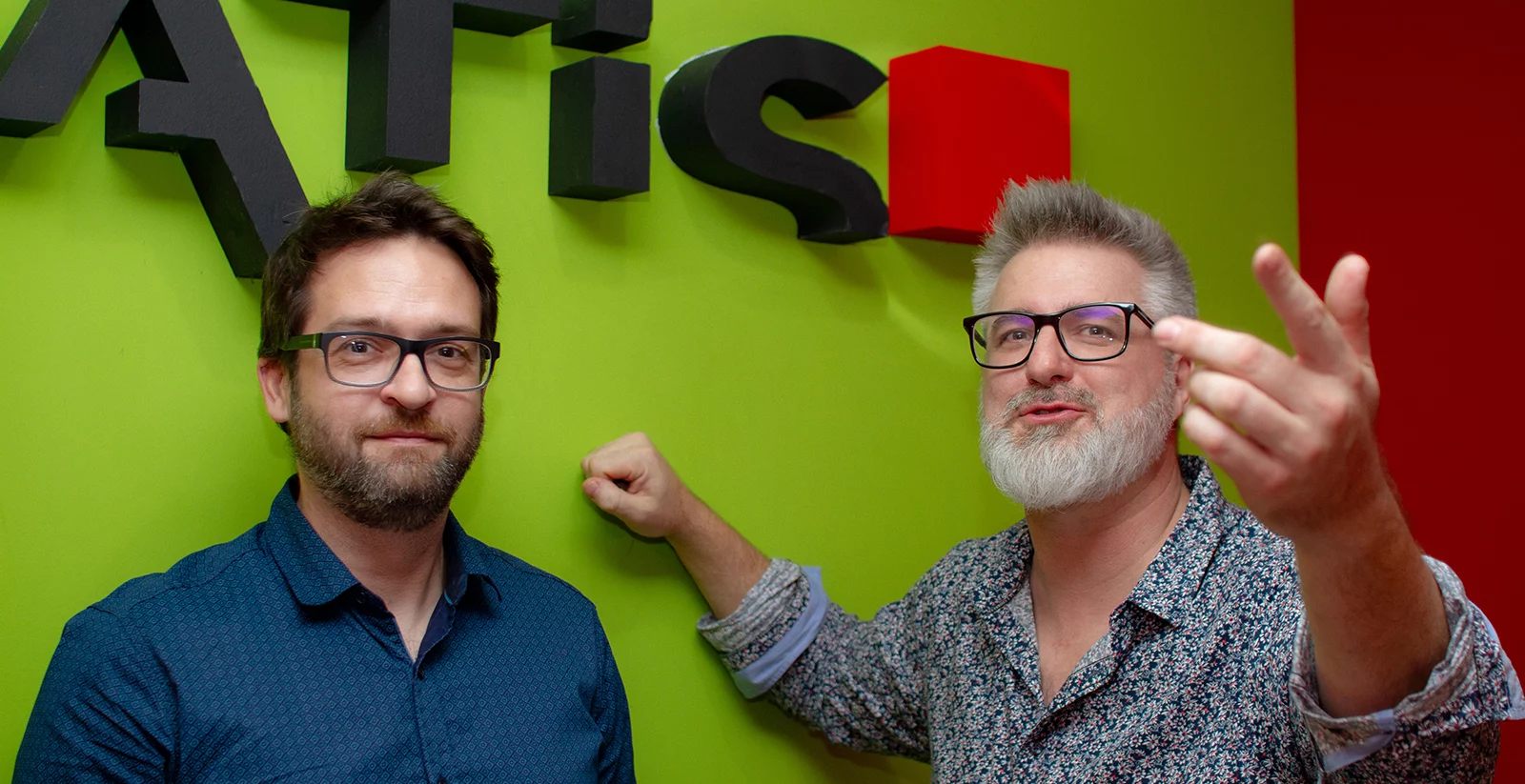 Deux hommes posent devant un mur vert avec une partie d’un logo en relief, comprenant des lettres noires et un cube rouge. L’homme de gauche porte des lunettes noires, une chemise bleue et a une barbe courte. Il a une expression neutre avec un léger sourire. L’homme de droite, avec une barbe blanche et des lunettes, porte une chemise à motifs et adopte une posture plus expressive, levant la main et semblant parler. L’arrière-plan coloré donne une ambiance dynamique à la scène.