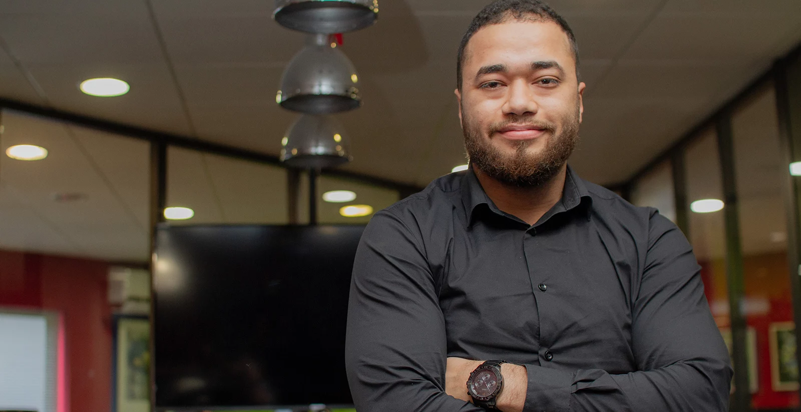 Un homme avec une barbe courte et des cheveux courts pose les bras croisés dans un bureau moderne. Il porte une chemise noire et une montre sombre à son poignet gauche. Son expression est confiante avec un léger sourire. L’arrière-plan montre un espace de travail avec un grand écran noir, des parois vitrées, des lumières suspendues et des murs aux tons rouges et verts, créant une ambiance professionnelle et dynamique