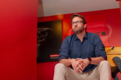 Un homme barbu portant des lunettes et une chemise bleu foncé est assis avec une posture décontractée, les mains croisées, dans une pièce aux murs rouges décorée d'affiches, dont l'une représente un visage de gorille dans une ambiance sombre. - Agrandir l'image 3 sur 12, fenêtre modale