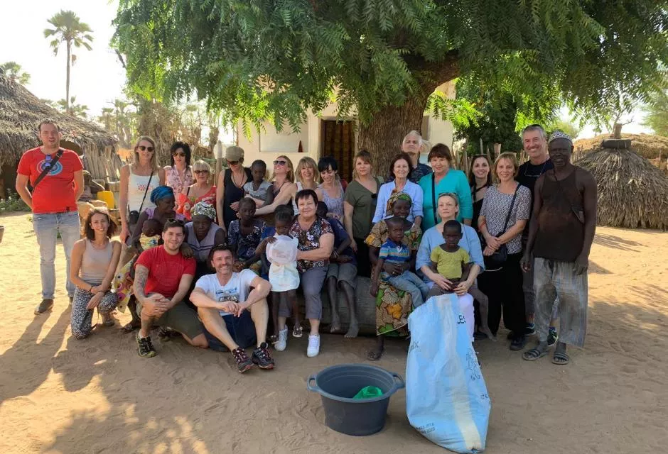 Un grand groupe de personnes, composé d’hommes et de femmes de différentes origines, pose sous un arbre imposant dans un village rural. Parmi eux, plusieurs adultes d'apparence européenne sont entourés d'habitants locaux, notamment des femmes et des enfants d'origine africaine. Certains membres du groupe sont assis sur un tronc d’arbre, d’autres sont debout. Les expressions varient entre sourires et regards bienveillants. À l’arrière-plan, on aperçoit des huttes en toit de chaume, un sol sablonneux et des palmiers, suggérant un cadre rural et chaud. Au premier plan, un grand sac en plastique et une bassine sont posés au sol, évoquant une action humanitaire .