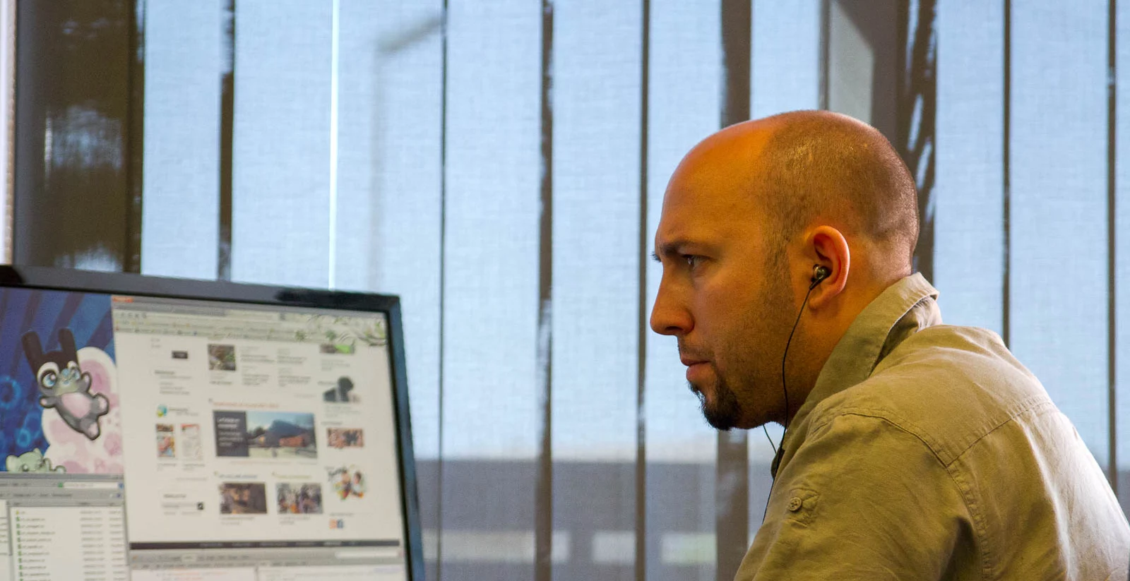 Un homme chauve avec une barbe courte est assis à son bureau, concentré sur l’écran de son ordinateur. Il porte une chemise beige et des écouteurs intra-auriculaires noirs. Son expression est sérieuse et attentive. L’écran affiche un navigateur web avec différentes pages ouvertes, ainsi qu’un fond d’écran illustré. Derrière lui, une grande baie vitrée avec des stores verticaux laisse entrer la lumière naturelle, offrant une vue partiellement floutée sur l’extérieur.