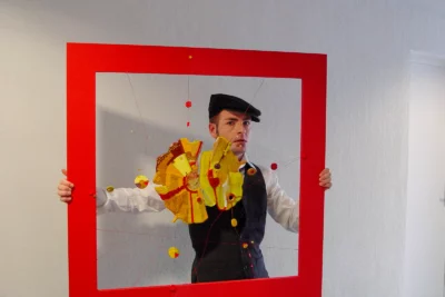 Un homme vêtu d’une tenue rétro avec un gilet noir, une chemise blanche et une casquette plate tient devant lui un grand cadre rouge contenant une composition artistique en fils et éléments colorés, créant un effet visuel original où il semble faire partie intégrante de l’œuvre. - Agrandir l'image 14 sur 28, fenêtre modale