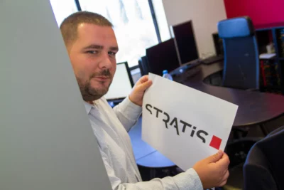 Un homme barbu vêtu d’une chemise blanche tient une feuille affichant le logo "STRATIS" en regardant vers l’objectif avec un sourire discret, debout dans un bureau équipé de plusieurs écrans et d’un grand bureau, avec des murs aux tons rouges et gris. - Agrandir l'image 6 sur 28, fenêtre modale