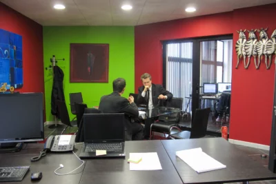 Deux hommes en costume sont assis à une table en verre dans un bureau aux murs rouges et verts, en pleine discussion sérieuse, tandis qu’à l’arrière-plan, un autre espace de travail avec plusieurs écrans d’ordinateur est visible, décoré de peintures colorées et de sculptures murales stylisés. - Agrandir l'image 12 sur 12, fenêtre modale