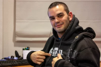 Un jeune homme souriant, aux cheveux courts et à la barbe légère, porte un sweat à capuche noir et pointe du doigt un motif imprimé sur son vêtement, assis devant un bureau avec un clavier et des accessoires informatiques en arrière-plan. - Agrandir l'image 5 sur 12, fenêtre modale