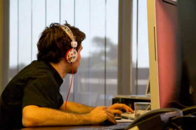 Un jeune homme concentré, vêtu d’une chemise noire et portant un casque audio décoré de motifs colorés, travaille sur un ordinateur dans un bureau baigné de lumière naturelle, les yeux fixés sur son écran et les mains sur le clavier et la souris. - Agrandir l'image 17 sur 28, fenêtre modale