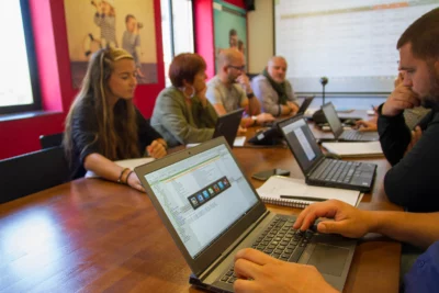 Un groupe de personnes est réuni autour d'une grande table de réunion, chacun travaillant sur un ordinateur portable ou prenant des notes, tandis qu'un grand écran projette un tableau de données dans une salle aux murs rouges décorée d'affiches. - Agrandir l'image 4 sur 12, fenêtre modale