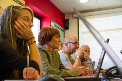 Un groupe de personnes assises autour d'une table dans une salle de réunion, concentrées sur une présentation projetée sur un écran, avec des expressions sérieuses et attentives. - Agrandir l'image 28 sur 28, fenêtre modale