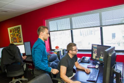 Dans un bureau aux murs rouges, un homme en chemise bleue se tient debout à côté d’un collègue en t-shirt noir assis devant un écran d’ordinateur affichant un logiciel de création graphique, tandis qu’un troisième employé travaille à son propre poste en arrière-plan. - Agrandir l'image 6 sur 12, fenêtre modale