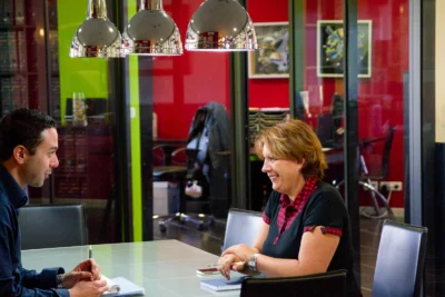 Un homme et une femme sont assis face à face à une table en verre dans une salle de réunion moderne, échangeant avec le sourire, sous des lampes chromées suspendues, avec des bureaux vitrés et des murs colorés en arrière-plan. - Agrandir l'image 7 sur 12, fenêtre modale