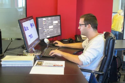 Un homme portant des lunettes et un t-shirt clair travaille sur un projet graphique devant deux écrans d’ordinateur, concentré sur son clavier dans un bureau aux murs rouges. - Agrandir l'image 18 sur 28, fenêtre modale