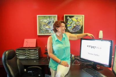 Une femme souriante, habillée en turquoise, est assise sur le bureau et pose un bras sur un écran d’ordinateur affichant le logo "STRATIS Marchés Publics", dans un bureau à la décoration colorée avec un mur rouge et des œuvres d'art modernes accrochées derrière elle. - Agrandir l'image 24 sur 28, fenêtre modale