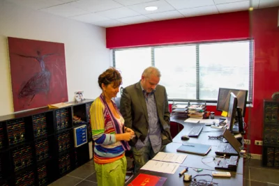 Un homme en costume gris et une femme vêtue d’un haut coloré et d’un pantalon vert se tiennent debout dans un bureau lumineux, examinant des documents posés sur un bureau bien organisé, dans une pièce décorée d’une grande peinture abstraite et de nombreuses étagères remplies de dossiers. - Agrandir l'image 5 sur 28, fenêtre modale