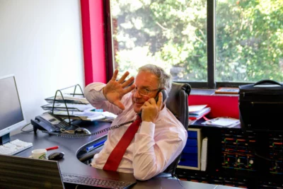 Un homme en chemise blanche et cravate rouge, assis à son bureau, parle au téléphone tout en levant la main avec un sourire, dans un bureau lumineux avec une grande fenêtre donnant sur un extérieur verdoyant. - Agrandir l'image 27 sur 28, fenêtre modale