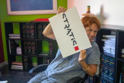 Une femme souriante aux cheveux courts roux est assise dans un bureau coloré et tient une affiche blanche avec le logo "STRATIS" écrit en noir avec un carré rouge à la fin. - Agrandir l'image 1 sur 12, fenêtre modale