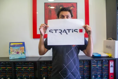 Un homme aux yeux expressifs et à l’air malicieux tient une feuille affichant le logo "STRATIS" devant son visage, ne laissant apparaître que ses yeux écarquillés. L’arrière-plan, soigneusement organisé, comprend une étagère avec des classeurs, une boîte marquée "Stratis" et une affiche encadrée en rouge, ajoutant une touche graphique et dynamique à l’image. - Agrandir l'image 21 sur 28, fenêtre modale