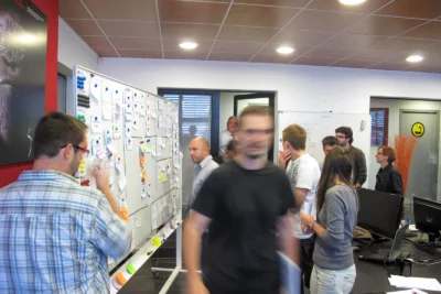 Un groupe de personnes est réuni dans un bureau dynamique, interagissant autour d’un grand tableau blanc couvert de notes et de post-it, avec certains en pleine discussion tandis que d’autres observent ou écrivent, dans une pièce moderne décorée d’affiches et d’éléments colorés. - Agrandir l'image 11 sur 12, fenêtre modale