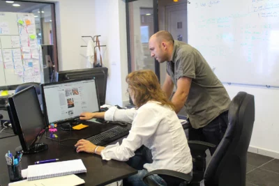 Une femme en chemise blanche est assise à son bureau, pointant son écran d’ordinateur, tandis qu’un homme en chemise kaki se penche à côté d’elle pour observer, dans un bureau moderne avec un tableau blanc rempli de notes et un mur couvert de post-it en arrière-plan. - Agrandir l'image 9 sur 12, fenêtre modale