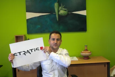 Un homme en chemise blanche, assis à son bureau, tient une feuille avec le logo "STRATIS" devant lui, sur fond de mur vert vif décoré d'une peinture abstraite et d'une étagère contenant des objets colorés. - Agrandir l'image 26 sur 28, fenêtre modale