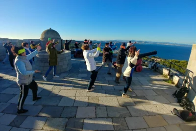 Un groupe de personnes participe à une séance de taï-chi en plein air sur une terrasse en pierre surplombant la mer, sous un ciel bleu éclatant, dans un cadre historique avec un ancien canon et un dôme en pierre en arrière-plan, alliant sérénité et dynamisme dans un cadre exceptionnel. - Agrandir l'image 10 sur 28, fenêtre modale