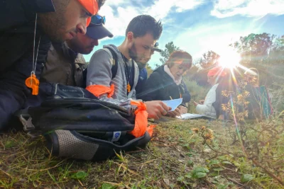 Un groupe de personnes, concentré et accroupi au sol en pleine nature, analyse une carte ou un document dans le cadre d’une activité de réflexion ou d’orientation, avec des bandanas et sifflets orange comme signes distinctifs, tandis que le soleil couchant crée un effet lumineux spectaculaire en arrière-plan. - Agrandir l'image 11 sur 28, fenêtre modale