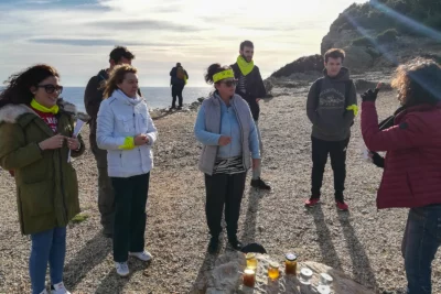 Un groupe de personnes, certaines portant des bandanas jaunes, est rassemblé sur une plage de galets face à la mer, écoutant attentivement une femme en doudoune rouge qui semble leur expliquer quelque chose, tandis que plusieurs pots de miel ou de confiture sont alignés sur un rocher sous un soleil éclatant. - Agrandir l'image 16 sur 28, fenêtre modale