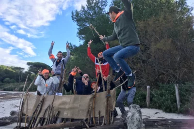 Un groupe de personnes participe avec enthousiasme à une activité en plein air sur une plage, rassemblées derrière une construction de bois et de toile de jute, faisant des signes de victoire et riant, tandis qu’un homme bondit en l’air dans une pose dynamique, ajoutant une touche humoristique et énergique à la scène. - Agrandir l'image 8 sur 28, fenêtre modale