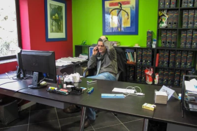 Un homme assis à son bureau, en pleine conversation téléphonique, passe une main dans ses cheveux d’un air concentré, entouré d’un espace de travail coloré avec des murs verts et rouges, des étagères remplies de classeurs et un bureau encombré de documents et fournitures. - Agrandir l'image 25 sur 28, fenêtre modale