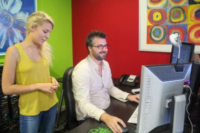 Un homme barbu portant des lunettes rouges et une chemise claire est assis à son bureau, souriant en travaillant sur son ordinateur, tandis qu’une femme blonde en débardeur jaune se tient à côté de lui, observant l’écran dans un bureau coloré aux murs rouges et verts, décoré de peintures artistiques. - Agrandir l'image 1 sur 28, fenêtre modale