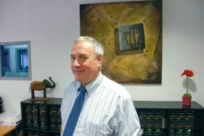 Un homme aux cheveux grisonnants, vêtu d'une chemise rayée et d'une cravate bleue, pose avec un léger sourire et un regard malicieux dans un bureau décoré d'une grande peinture abstraite, d'objets de décoration et de sculptures. - Agrandir l'image 10 sur 12, fenêtre modale