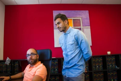 Un homme en chemise en jean se tient debout, observant un collègue barbu portant des lunettes et un polo orange, assis à son bureau et regardant vers l’objectif, dans un bureau aux murs rouges décoré d’une peinture abstraite et de nombreuses étagères remplies de dossiers. - Agrandir l'image 3 sur 28, fenêtre modale